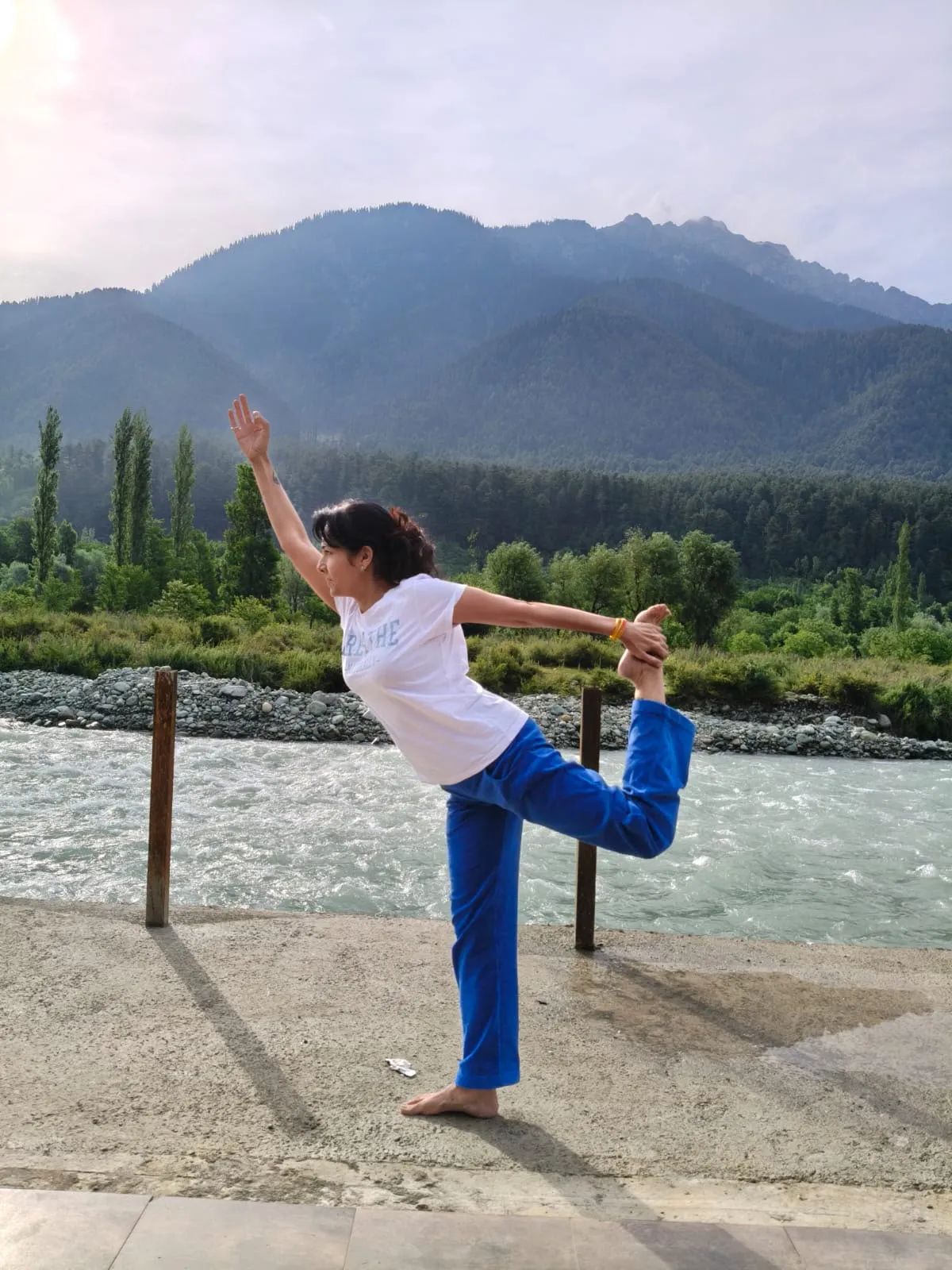 Vin in dancer’s pose on a riverbank, forested mountains behind
